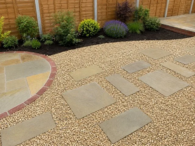 Golden gravel with square sandstone treads set through it, beside a circular sandstone feature ringed in red brick, with a planted border along a timber fence