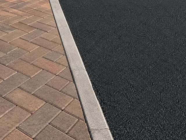 Concrete kerb running between fresh black tarmac and brindle block paving