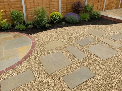 Golden gravel with square sandstone treads set through it, beside a circular sandstone feature ringed in red brick, with a planted border along a timber fence