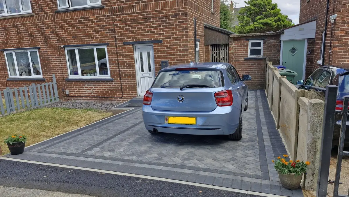 A light blue hatchback parked on a new grey herringbone block paved drive with charcoal borders, between a house and a fence, leading to a brick outbuilding