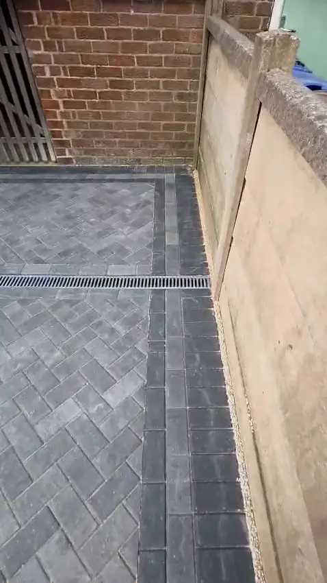 A steel channel drain set across the block paving at the far end of the drive, beside the fence
