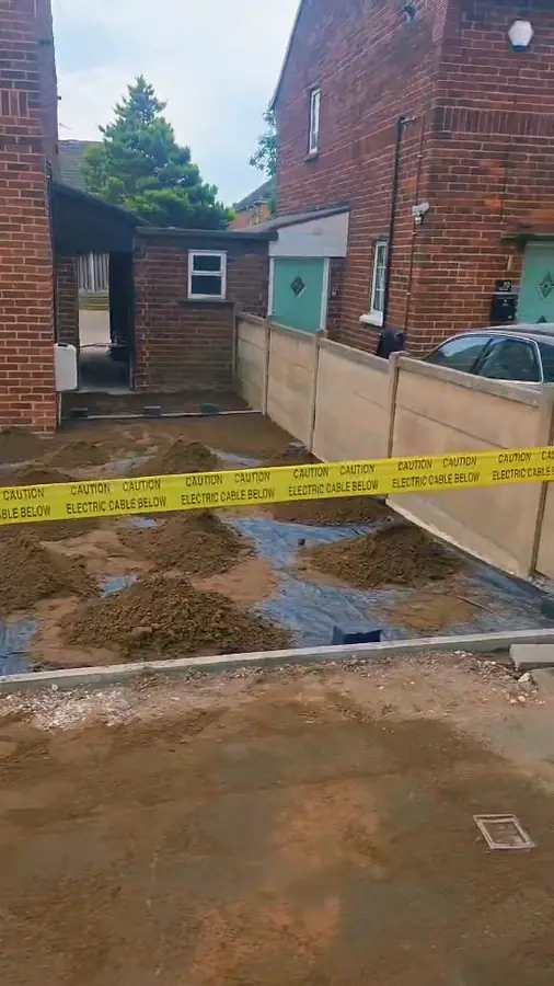 A dug-out drive beside a house, covered in black membrane with heaps of sand and a strip of yellow warning tape across it