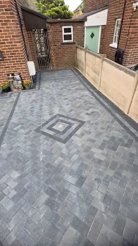 A diamond pattern in charcoal blocks set into grey herringbone block paving, with a charcoal border along a concrete fence