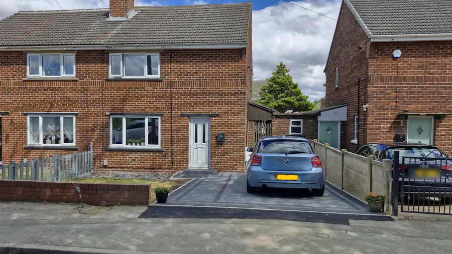 A semi-detached brick house with a new block paved drive in front of it, a light blue car parked on the drive and a concrete-post fence along one side