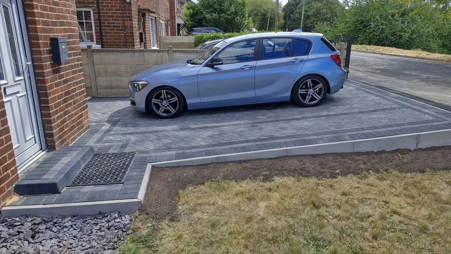A light blue car parked side-on on the new drive, beside a front door step with a mat set into the paving, a bed of slate chippings and a lawn edged by the border
