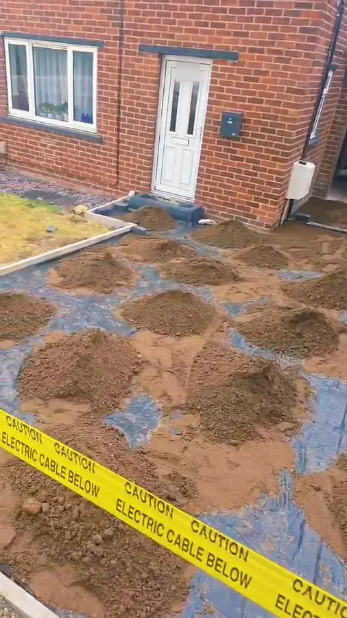 Heaps of sand on black membrane in front of a house, with yellow tape reading Caution electric cable below across the foreground